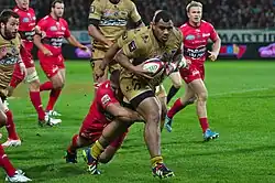 Joueur en rouge enserrant la cuisse gauche du porteur du ballon en maillot or. Derrière eux, des joueurs des deux équipes.