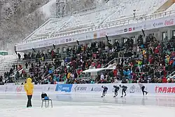 Un juge habillé en jaune se tient à côté d'une chaise. En arrière-plan, des patineurs concourent.