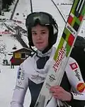 portrait tête et épaules de Urša Bogataj avec son casque et ses skis à la main, devant la piste de réception du tremplin de Brotterode