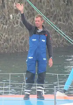Debout à l'avant de son bateau, le navigateur, en solopette bleue, salue la foule.