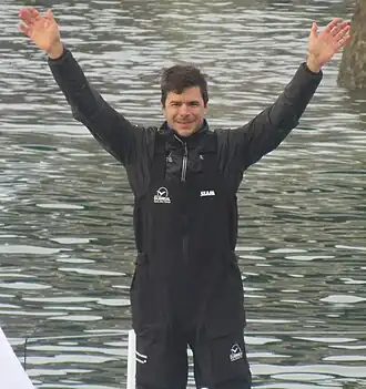 Le navigateur, de face, en buste, tout de noir vêtu, salue la foule des deux bras.