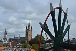 Photographie en couleurs d'un paysage urbain vu en hauteur, un clocher d'église pointant sur la gauche, une roue peinte en noir rouge et blanc et munie de trois axes visible sur la droite.