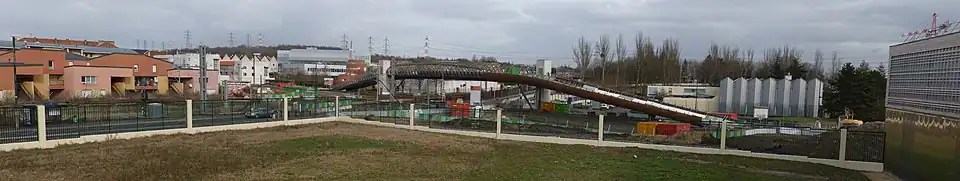 Le premier ouvrage d'art construit sur le T11 : la passerelle de Villetaneuse-Université, photographiée pendant son chantier de construction, desservant la gare construite dans un second temps à cheval sur les voies, et qui relie l'Université Paris-XIII, dont on voit le gymnase universitaire à droite du cliché, et le quartier du Centre-ville, à gauche. Au fond à droite, le Centre nautique Jacques-Duclos, démolit depuis.