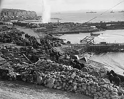 Photographie d'une plage jonchée de matériels abandonnés. Plusieurs attelages avancent sur un chemin le long de la mer où se trouvent deux pontons improvisés.
