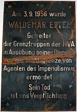 Plaque de métal endommagée indiquant « Am 3.9.1956 wurde WALDEMAR ESTEL Getreiter der Grenztruppen der NVA in Ausübung seine Dienstes an der Staatsgrenze von Agenten des Imperialismus ermordet. Sein Tod ist uns Verpflichtung. »