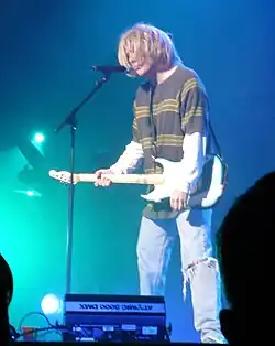 un homme habillé et coiffé comme Kurt Cobain jouant de la guitare sur une scène.