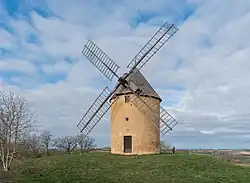 Photo du moulin de Gensac, un moulin avec une tour cylindrique en brique et pierre avec toiture en bardeau. Les ailes en bois ne sont pas équipées de toile. L'édifice se trouve sur une petite colline qui surplombe le paysage.