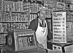 Photographie en noir et blanc d’un épicier souriant, cravaté, portant un tablier blanc à bretelle unique passant derrière le cou, coiffé d’un calot publicitaire ; l’homme s’appuie à une machine distributrice de café et à un comptoir surmonté de panneaux publicitaire ou annonçant les prix de marchandises. Derrière lui, l’étagère qui court tout le long du mur est remplie d’une multitude de boîtes de conserve, parfois cachées par des calicots publicitaires supplémentaires, donnant l’impression qu’il n’y a plus 1 cm2 de disponible.