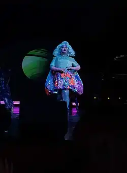 Kim Chi est sur scène. Son haut et sa perruque sont bleu-vert irisé ; sa jupe a un motif floral multicolore.