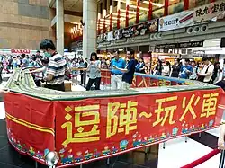 Plusieurs modélistes présentent une maquette de trains de firme circulaire. Ils sont au centre de la maquette pour la commander, le public étant face au décor, à l'extérieur du cercle.