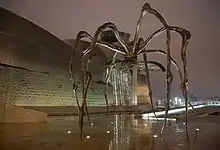 Sur le parvis d'un bâtiment d'architecture moderne, une gigantesque araignée de métal