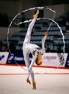 gymnaste debout sur un pied, penchée en arrière, dessinant des cercles avec un ruban. elle est vêtue d’une combinaison argentée recouvrant ses bras, son torse et ses jambes, décorée de noir à l’encolure
