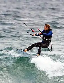 Photographie d'une femme sur une planche, tenant une barre accrochée par deux câbles à une voile.