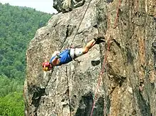 Au premier plan, une falaise sur la gauche avec au milieu un homme accroché à une corde. Il est horizontal avec sa tête légèrement incliné vers le bas ; ses pieds touchent la paroi. À l’arrière plan, une forêt.