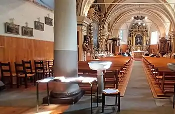 Photographie de l'intérieur d'une église avec les colonnes, les autels et les bancs ; on voit un maître-retable au fond de l'église ainsi que les différents ornements de l'édifice qui semblent baroques et gothiques au vu de leurs décorations opulentes pendant qu'une messe se déroule dans l'église en présence de fidèles.
