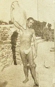 Photographie noir et blanc. Homme en maillot de bain portant sur son épaule une amphore.