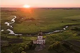 Photographie d'une plaine plate avec une rivière et un clocher d'église, sur fond de soleil couchant.