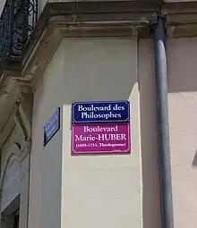 En dessous d'un panneau de rue bleu affichant "Boulevard des Philosophes", un second panneau violet affiche "Boulevard Marie-Huber,(1695-1753) Théologienne".