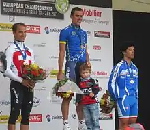Trois coureurs cyclistes debout sur un podium avec une médaille autour du cou et tenant un bouquet de fleurs.