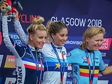 Trois femmes de face, plus ou moins jeunes, souriantes, toutes avec une médaille autour du cou, levant chacune un bras (le droit pour celles de gauche et du centre, le gauche pour celle de droite)