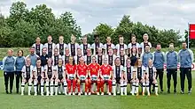 Photo de groupe de l'équipe d'Allemagne. Les joueuses sont photographiées en trois rangées, elles portent des maillots blancs avec une bande noire et leur numéros respectifs. Les gardiennes sont au centre au premier rang avec des maillot rouge portant des numéros jaunes. Le staff est situé de part et d'autre du rang central, avec des survêtements bleus.