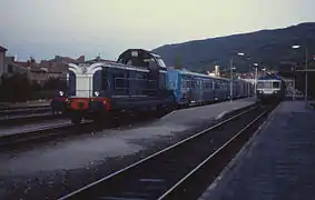 Photo couleur d'une locomotive diesel mono cabine bleu et blanche attelée à une rame bleu et inox à quai à gauche. À droite, un autorail bleu et blanc est de face.