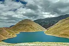 Petit lac aux eaux turquoises dans une cuvette montagneuse aux pentes enherbées sous un ciel grisâtre.