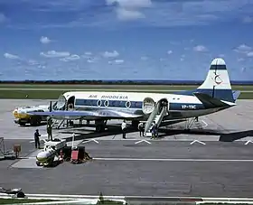 Un avion blanc et bleu sur une piste, entouré de camions-citernes de carburant et d'autres équipements. La queue porte un oiseau rouge stylisé du Zimbabwe, et les mots "AIR RHODESIA" sont peints au-dessus des fenêtres de la cabine. À l'arrière, les lettres "VP-YNC" sont peintes, surmontées du drapeau rhodésien vert et blanc.