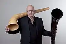 Armin Bachmann&nbsp;(de) posant avec deux parties de cor des Alpes, l'une en bois et l'autre en fibres de carbone.