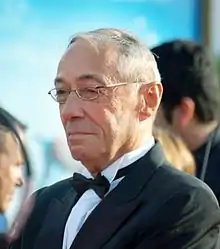 Plan poitrine d’un homme de 65 ans, cheveux gris courts, yeux fins et sombres, en smoking