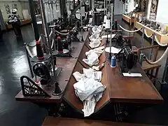 Photographie en vue rapprochée d'une rangée de postes de travail des ouvrières chemisières du XIXe&nbsp;siècle reconstitués au musée. Un unique meuble long en bois accueille deux rangées de places qui se font face, chacune pourvue d'une machine à coudre. Des séparateurs en métal arborant la marque Singer séparent les places. Au centre, une rigole creuse dans le bois accueille le linge. Chaque place dispose d'un tabouret.