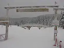 Clairière enneigée avec au premier plan un portail en bois et un insigne « Autrans : bienvenue à Gève, accès aux pistes », au second plan un ponton en bois et tout autour des conifères lourdement enneigés