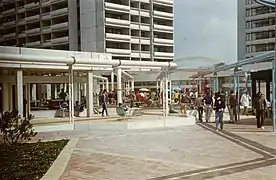 Photo d'une place du village olympique. On y distingue des canalisations blanches et bleues, et de nombreuses personnes se promenant. En fond, on voit un des grands immeubles.