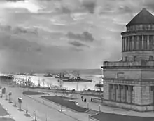 Photographie d'un grand monument de base cubique surmontée par une partie cylindrique à colonnades au sommet de laquelle se trouve un toit conique. Plusieurs cuirassés sont visibles dans le fleuve à l'arrière-plan.