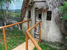 Façade d'une petite maison au mur blanc, avec une petite porte en bois, deux petits fenêtres de part et d'autre, et un toit en paille.