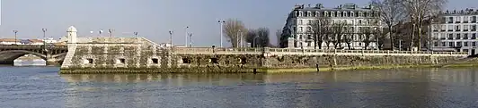 Vue d’un quai fortifié, orné d’une échauguette, en prolongement d’un pont sur un cours d’eau. À droite de la vue se dressent deux bâtiments imposants.