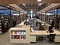 Vue lors de l'entrée dans la bibliothèque. On y voit le comptoir de prêt et les bornes de libre-service au centre ainsi que les documentaires adultes de chaque côté.