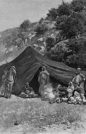 Groupe d'hommes assis et debout devant une tente, l'un d'eux, assis, tire un coup de fusil.