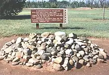 Un tas de pierres grossières, d'environ 3 m de diamètre et 40 cm de haut ; à l'arrière-plan, un panneau explicatif.