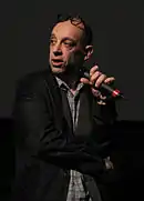 Un homme debout, presque chauve, croise les bras devant lui tout en tenant un micro, et regarde sur sa droite.