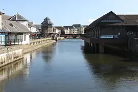La rivière canalisée et entourée de bâtiments, dont un marché à droite et une tour à horloge à gauche.