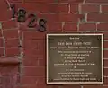 Photographie d'une plaque en métal sombre, avec des lettres claires, comportant un texte en anglais sur plusieurs lignes, sur un mur de briques rouges ; en haut à gauche, les chiffres 1828 en cascade.