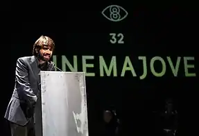 Homme souriant, debout derrière un pupitre, devant un fond noir avec un logo vert du festival