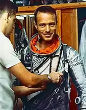 Un homme (de dos) en aide un autre (de face) à enfiler une combinaison spatiale.