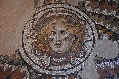 Dans un cercle, la tête de Méduse est enlacée par un couple de serpents qui se rejoignent sur la tête et un autre qui jaillissent des oreilles vers la base du nez.