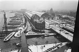 La gare centrale, vue depuis le nord-ouest, un jour de neige. Elle est bordée, à gauche par un terminal de bacs. Au premier plan, le Westertoegang&nbsp;(nl), court canal réunissant le lac IJ, à gauche, et le canal Singel, à droite. Entre les deux grandes verrières de la gare, il existe encore une partie à ciel ouvert