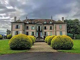 photographie en couleurs d'un bâtiment de teinte rose aux volets bleus devant une esplanade entourée de buis en boules.