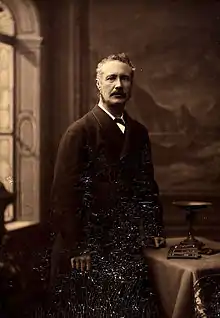 photographie sépia : portrait en pied d'homme moustachu appuyé sur une table
