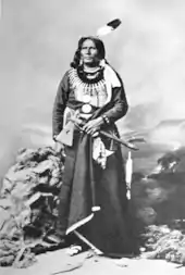 Photographie en noir et blanc d'un Amérindien en habit traditionnel coiffé d'une plume et tenant un tomahawk.