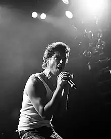Photographie en noir et blanc d'un homme debout sur scène chantant dans un micro.